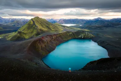 A tiny hiker by Lake Bláhylur in Landmannalaugar at sunset, surrounded by mountains in shades of green, ochre, and rhyolite—colors characteristic of the Icelandic Highlands.